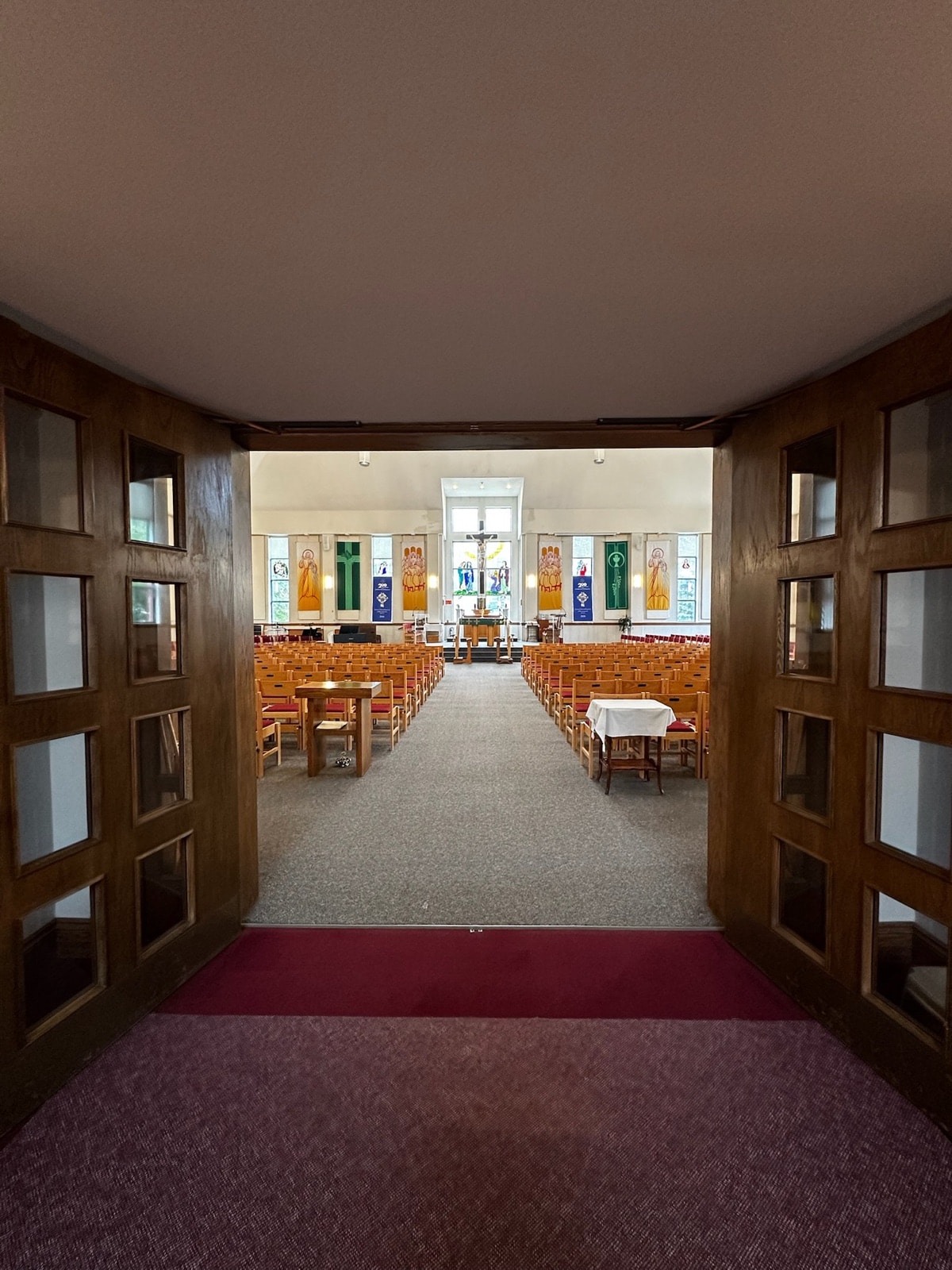 View through open wooden doors down a central carpeted aisle toward the altar, with rows of seating on both sides and a crucifix window at the far end.