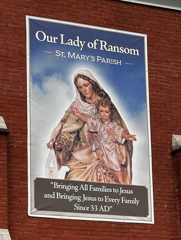 The parish's Our Lady of Ransom banner on the brick face of the entrance tower: Mary in a pale veil holding the child Jesus, above the words “Bringing All Families to Jesus and Bringing Jesus to Every Family Since 33 AD”.