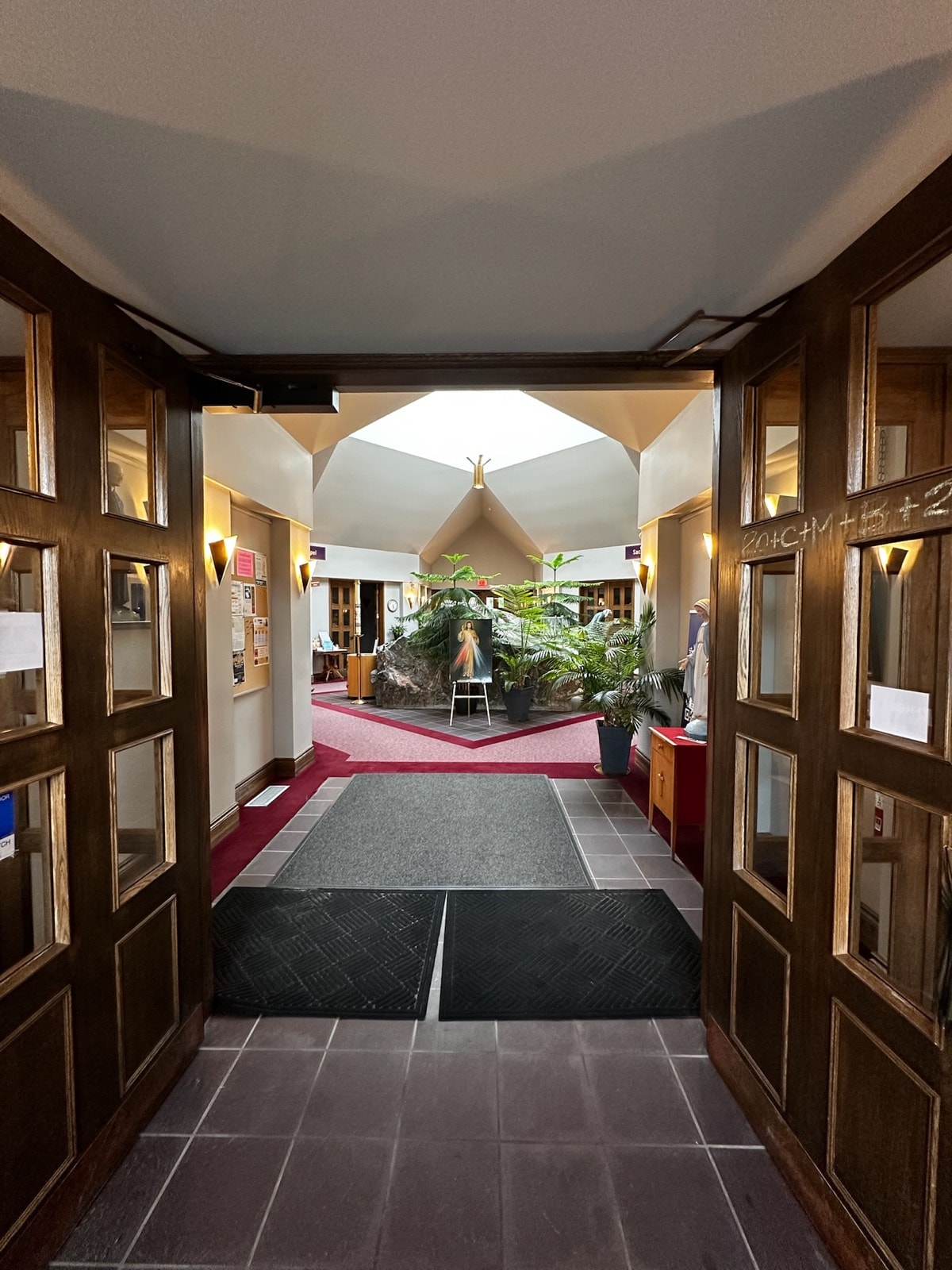 View through open wooden double doors into a naturally lit lobby with a rock feature, potted plants and a framed image on a stand.